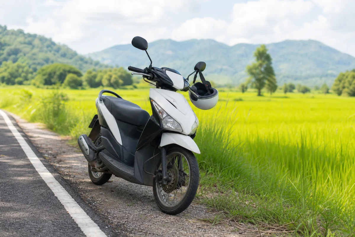 Scooter garé avec un casque accroché sur le guidon devant une route de campagne thaïlandaise
